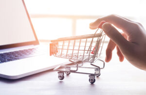 A small, metal shopping cart is pushed by a hand beside a laptop, symbolizing a digital purchase.